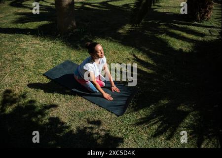 Femme pratiquant le yoga seule à l'extérieur par une journée ensoleillée, agenouillée sur une natte dans un parc verdoyant luxuriant, profitant de la tranquillité. Banque D'Images