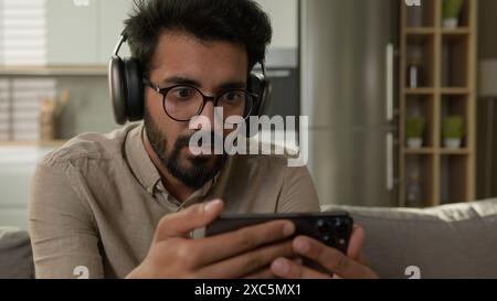 Choqué étonné homme arabe indien regarder la vidéo streaming sport en ligne match addict des médias sociaux homme concentré dans les écouteurs regardant le téléphone mobile Banque D'Images