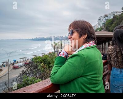 Lima, Pérou - juillet 30 2023 : une femme péruvienne portant un manteau vert se penche sur un balcon rouge et regarde la mer Banque D'Images