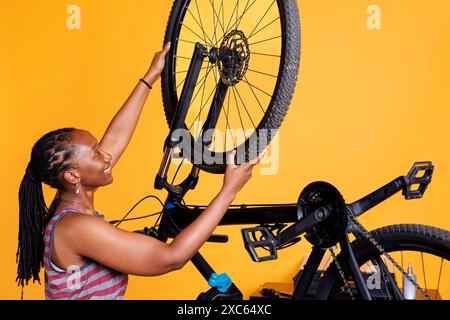 Femme noire amateur de sport examine attentivement un vélo, remplaçant un pneu arrière endommagé. Cycliste afro-américaine engagée en bonne santé rattachant en toute sécurité la roue de vélo devant le fond jaune. Banque D'Images