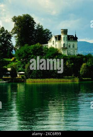 Une grande maison blanche se trouve sur une colline surplombant un plan d'eau. La maison est entourée d'arbres et l'eau est calme et claire. Concept de tranquili Banque D'Images