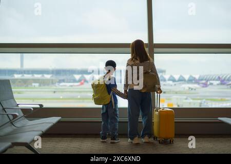 Mère asiatique et son jeune fils debout côte à côte à une fenêtre de l'aéroport, regardant tous les deux les avions sur le tarmac Banque D'Images