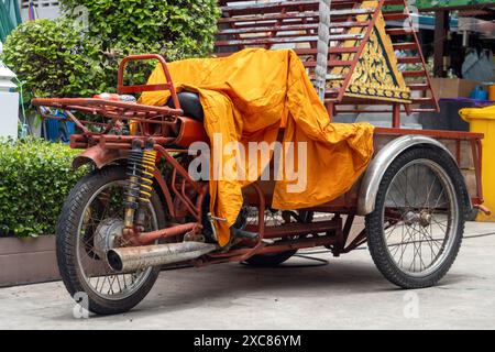 Un tricycle motorisé avec un chariot est couvert par les robes orange d'un moine bouddhiste, Thaïlande Banque D'Images