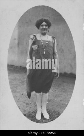 Une carte postale photographique en noir et blanc des années 1920 mettant en vedette un acteur amateur dans Drag. Le sujet, vêtu d'une robe à rabat et de chaussures blanches, se dresse contre un mur en toile de fond. Banque D'Images