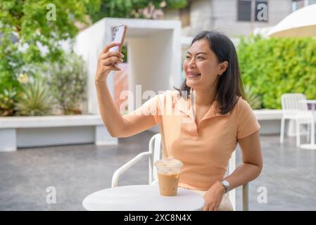 Femme assise dans un café en plein air. Elle est vêtue décontractée d'un dessus orange et apprécie un café glacé placé sur la table devant elle. Le s Banque D'Images