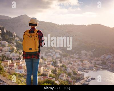 Nice asiatique heureuse femelle avec sac à dos profitant de ses vacances sur les îles Symi au coucher du soleil. Vue sur le port Symi ou Simi, est minuscule île du Dodécanèse, Grèce, atmosphère calme et architecture fabuleuse. Banque D'Images