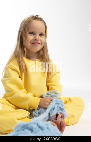 Jolie petite fille en vêtements jaunes joue avec boule de fil bleu. Modèle enfant avec de longs cheveux blonds assis dans le studio sur fond clair. Photo de haute qualité Banque D'Images