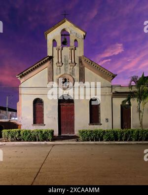 395 coucher de soleil sur Calle 2 de Diciembre Street et l'église Iglesia San Francisco Javier tourne le ciel d'une teinte violacée-rosée-rougeâtre. Niquero-Cuba. Banque D'Images