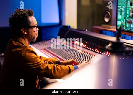 Expert audio traitant la musique à partir de la cabine vocale insonorisée dans un studio professionnel, enregistrant des chansons et les montant après la post-production. Le technicien utilise la console de mixage et les boutons ou curseurs. Banque D'Images