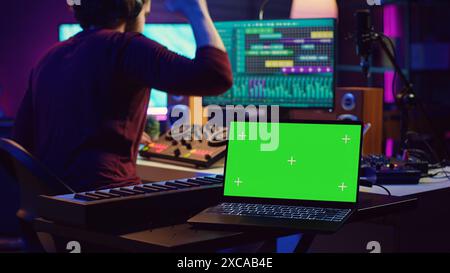 Ingénieur musical éditant des enregistrements audio sur console de mixage et logiciel daw, ayant un ordinateur portable avec écran vert dans home studio. Artiste produisant une nouvelle chanson avec des outils d'ingénierie acoustique. Caméra A. Banque D'Images