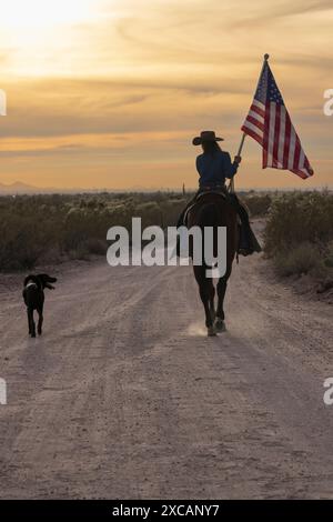 Main de ranch femelle, ou cowgirl, à cheval et portant le drapeau américain dans le coucher du soleil sur la route de terre sur un ranch en Arizona, États-Unis. Accompagné d'un chien. Banque D'Images