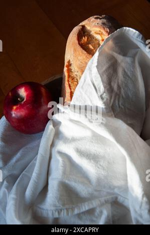 Une miche de pain frais enveloppée dans un linge de lin blanc avec une pomme rouge vif sur une table de ferme en bois Banque D'Images