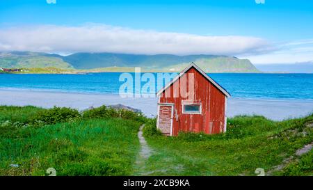 Une cabane rouge altérée se trouve sur une plage de sable blanc en Norvège, avec une mer turquoise et une chaîne de montagnes vertes au loin. Ramberg Beach Lofoten Norvège Banque D'Images