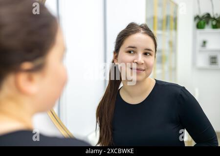 une fille regarde dans le miroir après la procédure de dermabrasion et est très heureuse qu'elle ait utilisé ce cosmétique. Une très belle petite fille portant un Banque D'Images