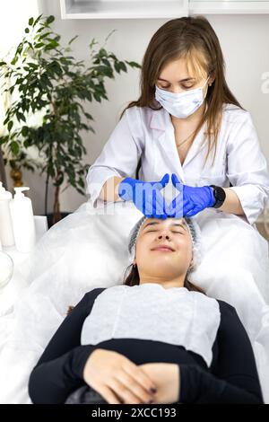 Cosmétologue faisant le massage du visage avec ses mains dans des gants en caoutchouc, au client dans la clinique de beauté. Le client est allongé o une table de traitement relaxant Banque D'Images