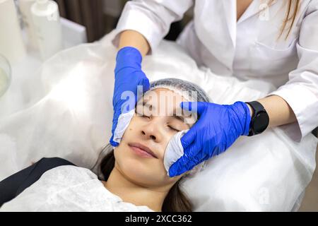 Dermatologue dans les gants en nitrile enlevant les restes de gel du visage de la jeune femme à l'aide de mouchoirs en papier, vue de dessus gros plan. Banque D'Images