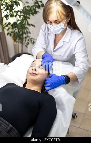 Esthéticienne portant des gants bleus en nitrile, mettant une coiffe sur la tête de son client pour se préparer à une intervention cosmétique. Banque D'Images