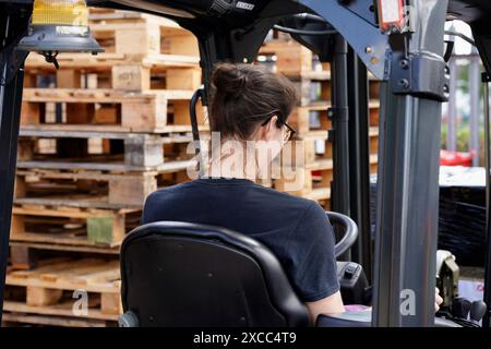 Jeune femme utilisant un chariot élévateur dans un entrepôt avec des palettes en bois en arrière-plan Banque D'Images