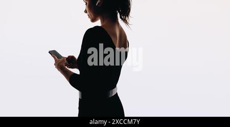 Femme se tient dans un studio, tenant un smartphone et portant des écouteurs. Elle tape sur une application mobile, s'engageant dans la communication sur les médias sociaux. Banque D'Images