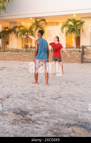 Deux jeunes hommes jouant au Frisbee sur une plage de sable en face d'un bâtiment moderne avec des palmiers. Banque D'Images