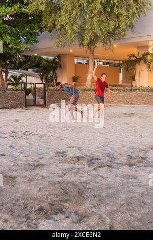 Deux jeunes hommes jouant au Frisbee sur une plage de sable au coucher du soleil avec un bâtiment moderne et des palmiers en arrière-plan. Banque D'Images