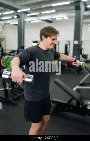 Plan vertical d'un sportif débutant motivé faisant des relèvements latéraux latéraux avec des haltères au gymnase. Homme beau actif s'exerçant pour rester en forme et mince Banque D'Images