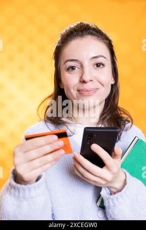 Femme avec pile de livres dans les mains excité d'acheter plus en utilisant la carte de crédit, isolé sur fond de studio. Bookworm tenant pile de romans et téléphone portable, achetant des trucs à lire Banque D'Images