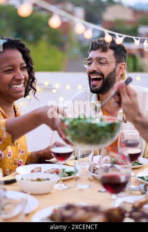 Femme afro-américaine souriante assise à la table en passant salade à des amis heureux à l'événement de repas en plein air Banque D'Images
