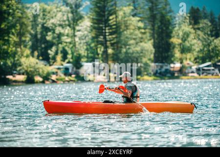 Un homme dans un gilet de sauvetage pagaie un kayak rouge sur un lac calme. L'eau est pétillante et le lac est entouré d'une forêt de grands arbres. Il y a un Banque D'Images