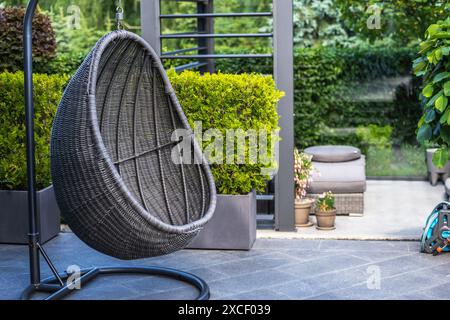 Un fauteuil à oeufs en osier noir est suspendu à un support en métal sur un patio entouré de verdure. La chaise est suspendue devant un grand buisson en pot, avec un gr Banque D'Images