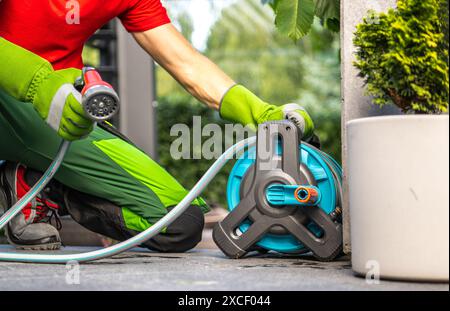 Un homme en pantalon vert et chemise rouge arrose les plantes à l'aide d'un tuyau et d'un enrouleur de tuyau. Il est agenouillé sur un patio, tenant la buse et dirigeant l'eau Banque D'Images