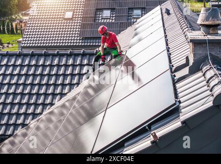 Un travailleur portant un casque rouge et un pantalon vert installe des panneaux solaires sur un toit résidentiel. Le toit est carrelé et les panneaux sont disposés en rangées. Banque D'Images