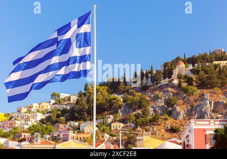 Drapeau grec ondulant avec des maisons colorées à flanc de colline et des arbres à Symi, Grèce sous un ciel bleu clair Banque D'Images