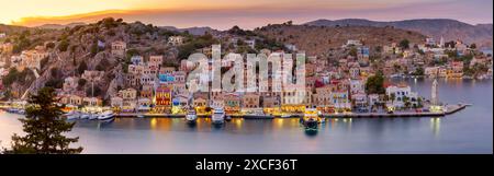 Vue panoramique du port de Symi au coucher du soleil avec des bâtiments et des bateaux illuminés, Grèce Banque D'Images