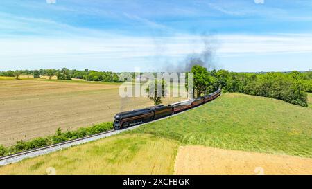 Vue aérienne captivante d'un train à vapeur vintage voyageant à travers un vaste paysage rural, avec des champs labourés et verts sous un ciel bleu. Banque D'Images