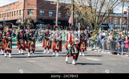 Lindenhurst, New York, États-Unis, le 30 mars 2024 - Un groupe de cornemuse habillés de kilts écossais traditionnels et de chapeaux en peau de Bearskin se produit dans Une Street Parade colorée, entourée de foules de spectateurs un jour ensoleillé dans un cadre urbain animé. Banque D'Images