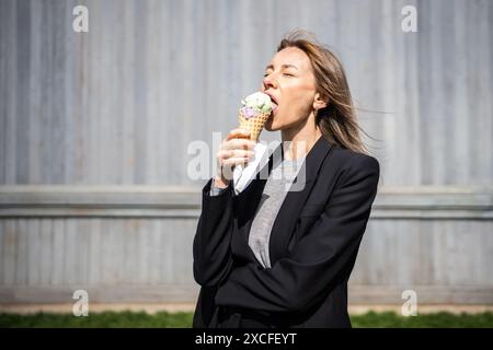 Entrepreneur de femme blonde détendue satisfaite appréciant la crème glacée dans le cône de gaufre prend pause à l'extérieur. Banque D'Images