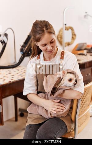 Portrait vertical de jeune femme souriante comme toiletteur de chien femelle tenant mignon petit chiot enveloppé dans une serviette pendant les soins de beauté Banque D'Images