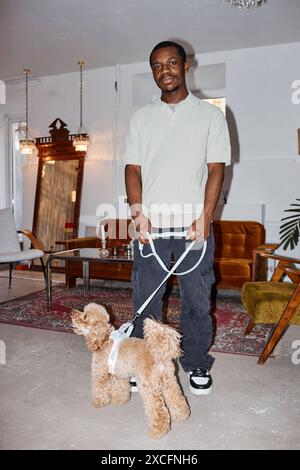 Portrait vertical pleine longueur de l'homme afro-américain tenant le chien mignon en laisse et regardant la caméra dans le studio de toilettage, tourné avec flash Banque D'Images