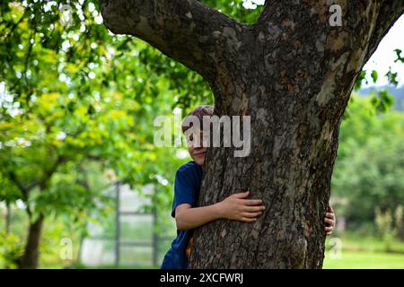 Le jeune garçon embrasse un grand arbre dans un parc, montrant son amour pour la nature. Banque D'Images