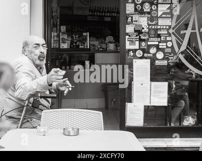 Cremona, Italie - 10 juin 2024 homme senior assis à une table de café en plein air, fumant une cigarette et faisant des gestes tout en parlant Banque D'Images