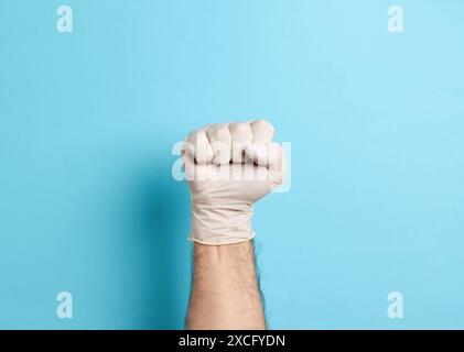 Main masculine portant un gant médical en latex blanc faisant un poing, sur un fond bleu Banque D'Images