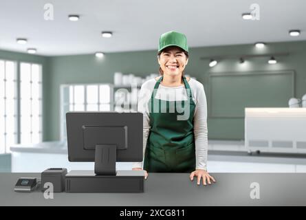 Le vendeur asiatique actif porte un tablier vert et un chapeau vert souriant avec le terminal de point de vente ou le bureau de caisse Banque D'Images