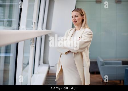 Belle femme blonde enceinte confiante dans des vêtements élégants et à la mode, portant une robe blanche et un blazer, debout avec les bras croisés dans une entreprise Banque D'Images