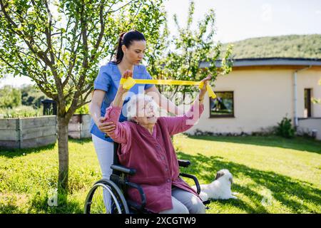 Soignant féminin faisant des exercices motorisés avec une femme âgée en fauteuil roulant. Infirmière et femme âgée travaillant avec bande de résistance. Banque D'Images