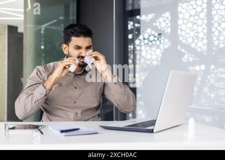 Homme éternuant devant l'ordinateur portable tout en travaillant au bureau, en utilisant des mouchoirs en raison du froid ou des allergies Banque D'Images