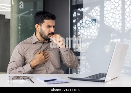 Homme toussant tout en travaillant sur un ordinateur portable à un bureau, tenant sa poitrine dans l'inconfort. Montre des signes de maladie ou de problèmes respiratoires. Banque D'Images