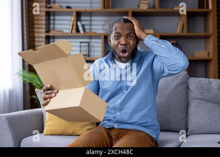 Homme surpris assis sur un canapé à la maison tenant une boîte vide avec une expression choquée sur son visage. Banque D'Images