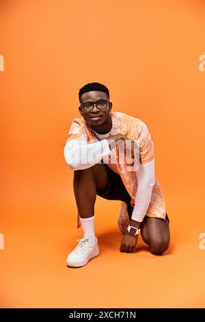 Jeune homme afro-américain à la mode accroupi dans des lunettes élégantes sur fond orange vif. Banque D'Images