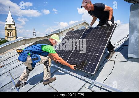 Ouvriers construisant un système de panneaux solaires sur un toit en métal en milieu urbain. Deux installateurs hommes transportant le module solaire photovoltaïque à l'extérieur. Concept de production d'énergie alternative, verte et renouvelable. Banque D'Images